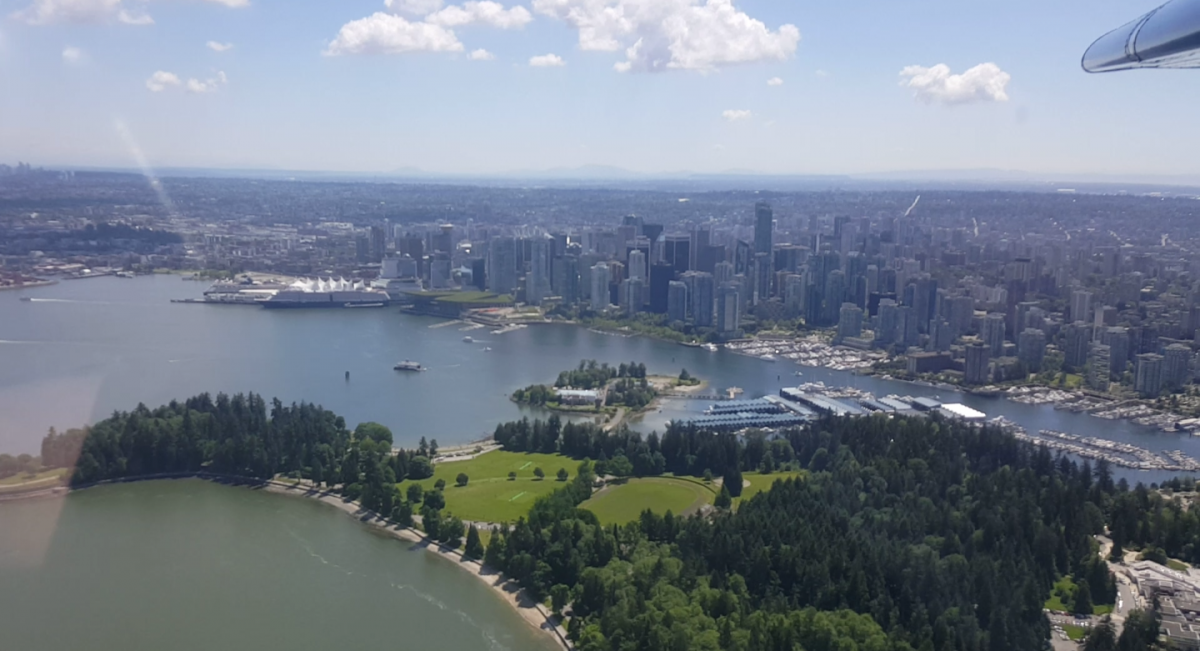 Foto aus dem Fenster eines Wasserflugzeugs, unten ist eine Küstenlandschaft mit gruenen Wiesen udn Baumen im Vordergrund zu sehen, dahinter die Skyline von Vancouver