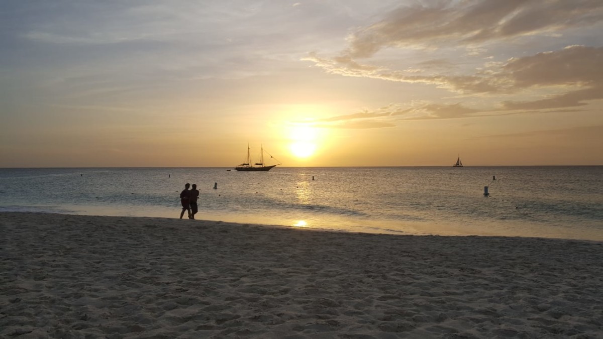 Mensch steht bei Sonnenuntergang am Strand, dahinter spiegelt sich die orange verfaerbte Sonne im Meer