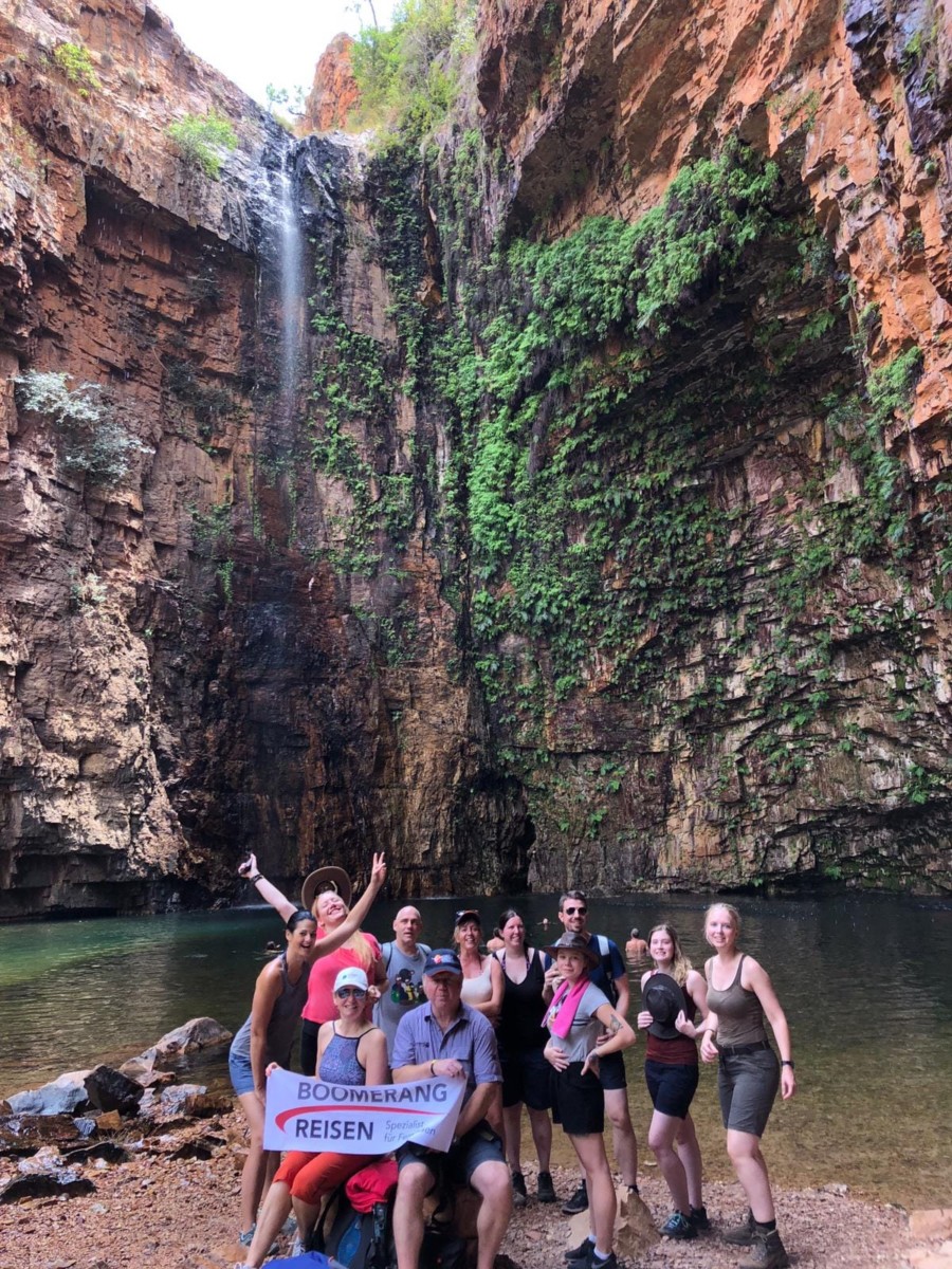 Eine Reisegruppe steht in einer Schlucht vor einem Wasserfall, der in einen See stürzt und könnt eine Flagge mit Boomerang Reisen in die Höhe