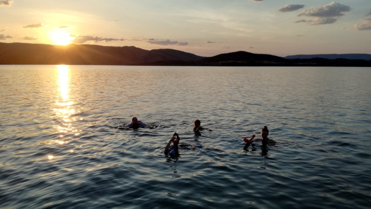 Vier Menschen schwimmen ausgelassen in einem See, im Hintergrund geht die Sonne über einer Bergkette unter