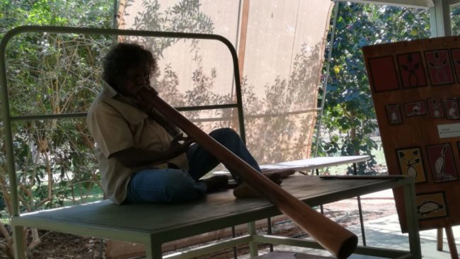 Aborigine sitzt auf einer Pritsche und spielt Didgeridoo