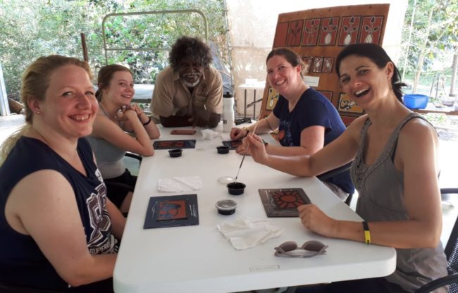 vier Menschen sitzen an einem Tisch und probieren sich unter Anleitung eines Aborigines im Dot Painting