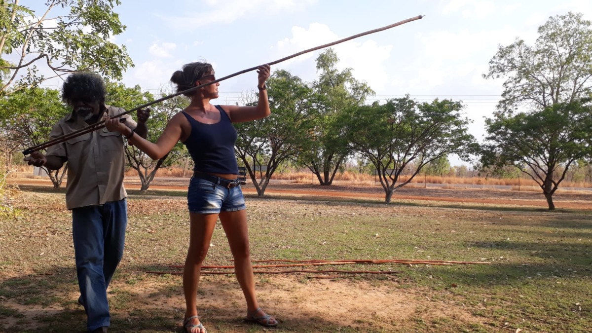 Frau bekommt auf einer freien Grasfläche mit wenigen Bäumen im Hintergrund von einem Aborigine die Wurftechnik beim Speerwerfen gezeigt,