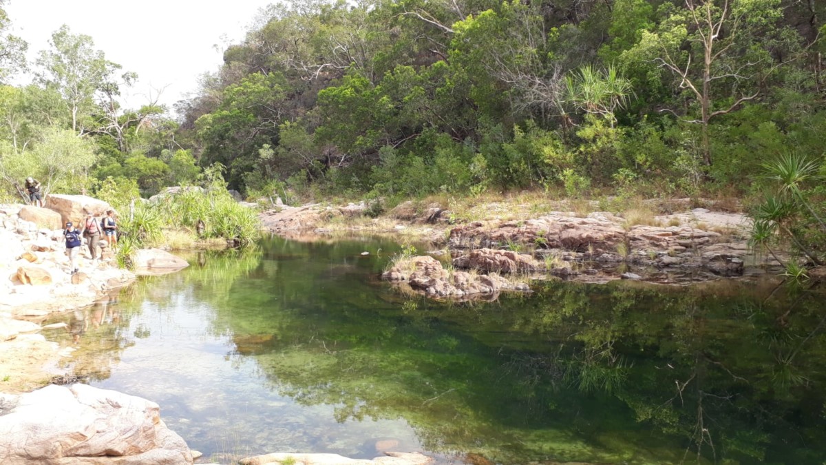 grüne Buschlandschaft spiegelt sich im Wasser einer Lagune wider, die von Felsen umgeben wird, vier Menschen klettern entlang des felsigen Ufers