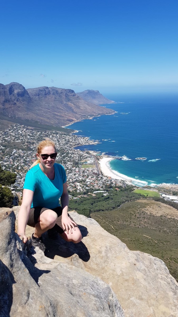 Frau sitzt auf einem Felsvorsprung, darunter sieht man Kapstadt, das Meer und den Tafelberg