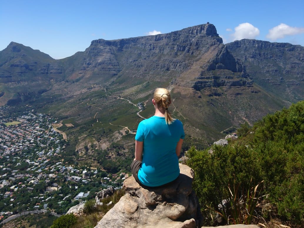 Frau sitzt auf einem Felsvorsprung und blickt auf die Landschaft mit Kapstadt im Tal und dem Tafelberg im Hintergrund