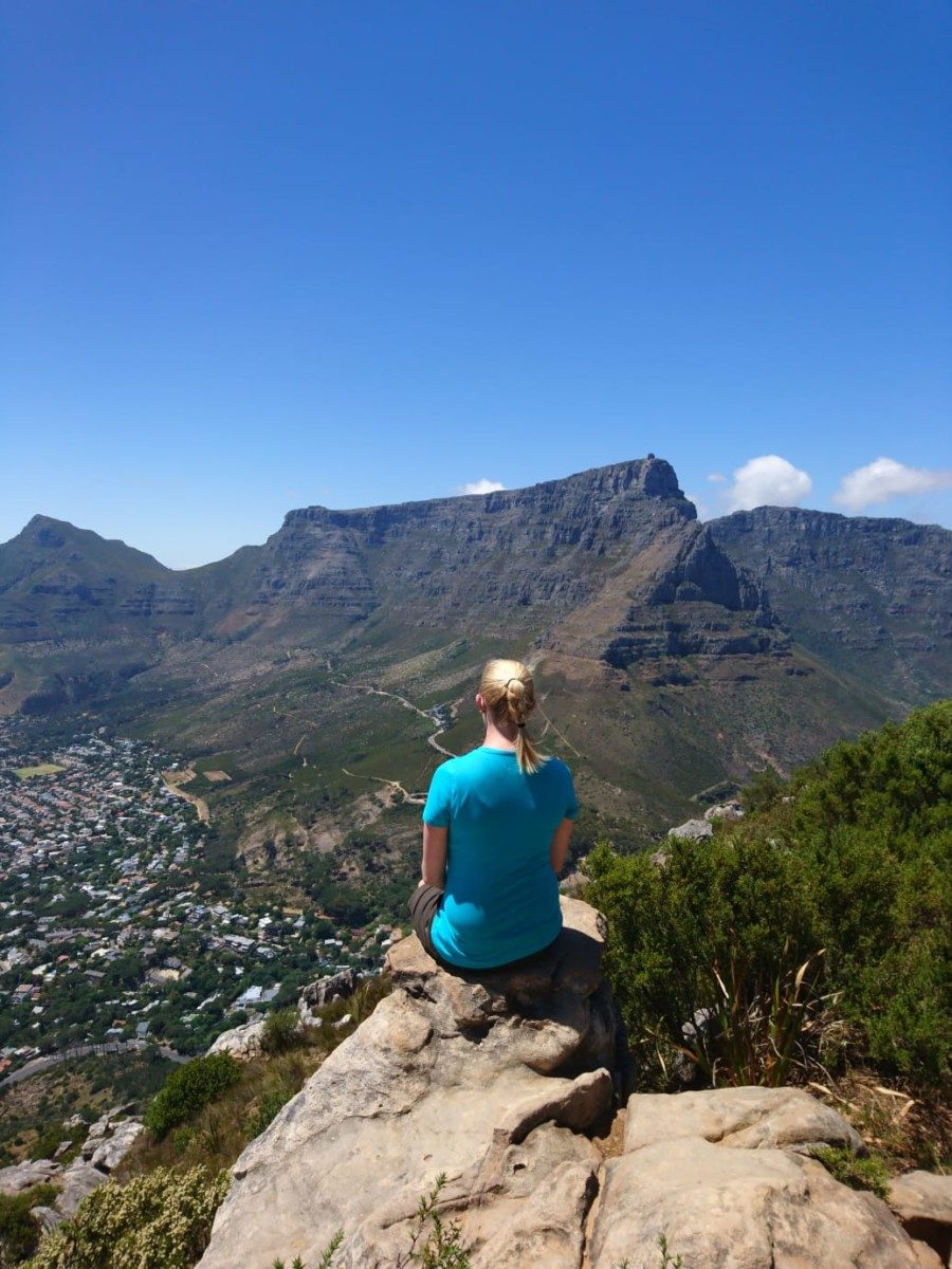 Frau sitzt auf einem Felsvorsprung und blickt auf die Landschaft mit Kapstadt im Tal und dem Tafelberg im Hintergrund